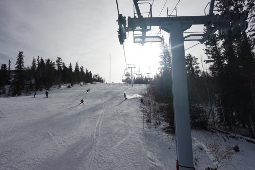 Skiers on a snowy slope with a ski lift in the foreground and trees in the background under a cloudy sky.