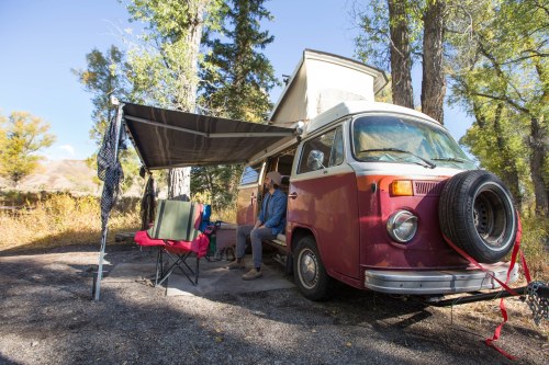 A vintage camper van parked under trees, with a person sitting outside on a chair, enjoying the outdoors.