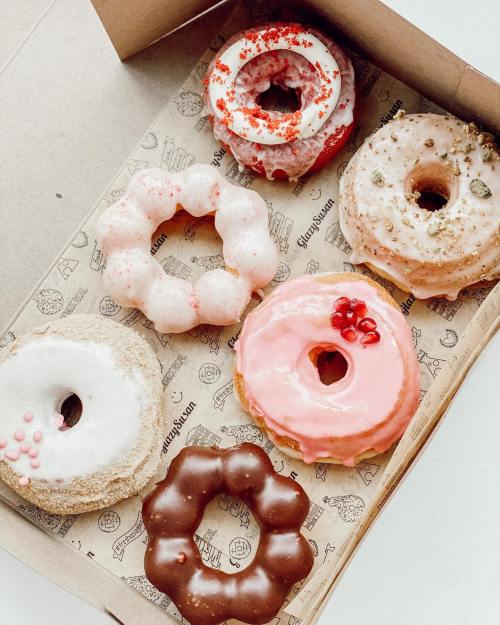 A box of six assorted donuts with various glazes and toppings, including pink, white, and chocolate varieties.