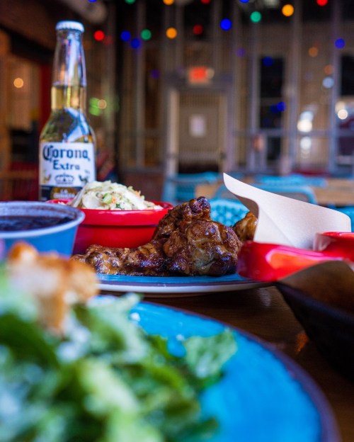 A vibrant table setting featuring a plate of chicken, sides, and a bottle of Corona, with colorful lights in the background.