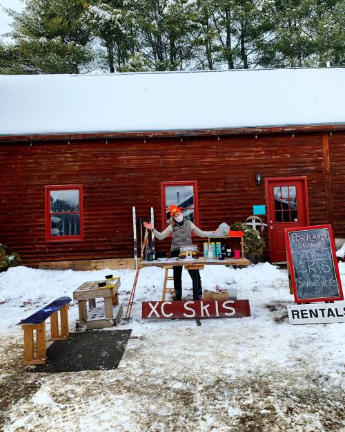 A person stands outside a rustic red building with skis, rental signs, and snow, welcoming visitors to a ski shop.