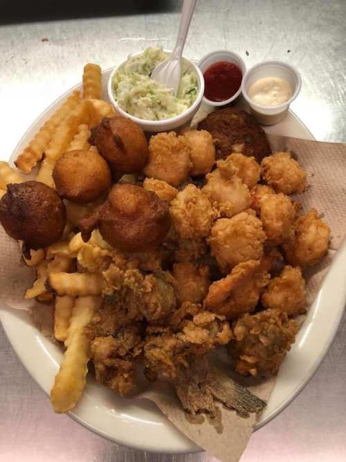 A plate of fried seafood, including shrimp, fish, hushpuppies, crinkle fries, and coleslaw with dipping sauces.