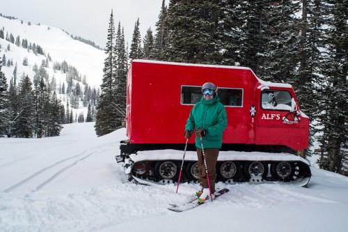 A skier in a green jacket stands next to a red snowcat vehicle in a snowy forested area.