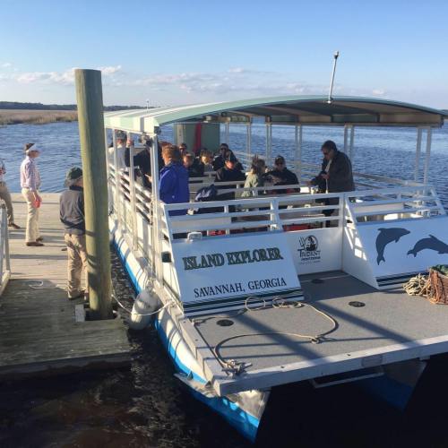 A group of people boarding the "Island Explorer" boat at a dock in Savannah, Georgia, with water and marshland in the background.
