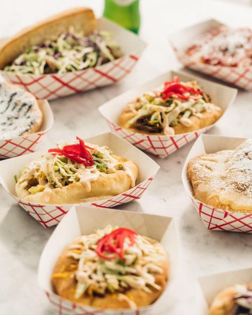 A variety of colorful hot dogs and sandwiches in paper trays, topped with vegetables and sauces, on a marble surface.