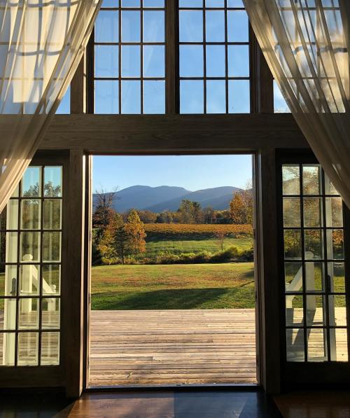 A scenic view of mountains and autumn foliage through large glass doors, framed by sheer curtains.