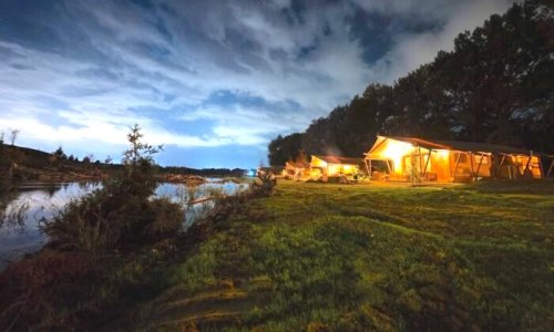 A serene campsite by a river at night, with glowing tents and a cloudy sky reflecting soft light.