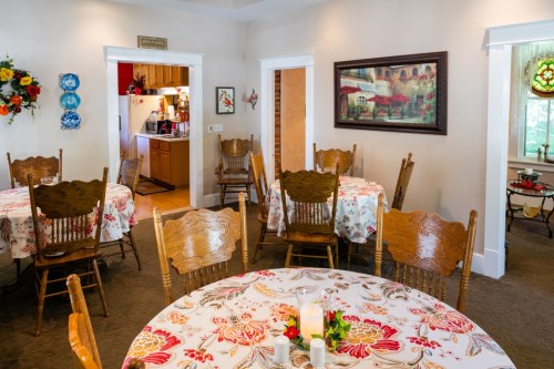 Cozy dining area with floral tablecloths, wooden chairs, and a view into a kitchen. Bright and inviting atmosphere.