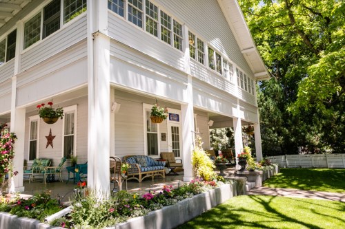 A charming white house with a porch, surrounded by colorful flowers and greenery on a sunny day.
