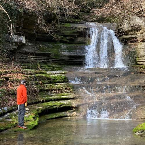 A person in an orange jacket stands by a mossy waterfall, surrounded by rocky terrain and trees.