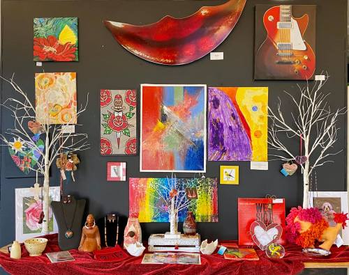 A colorful art display featuring paintings, sculptures, and decorative elements on a dark wall with a red tablecloth.