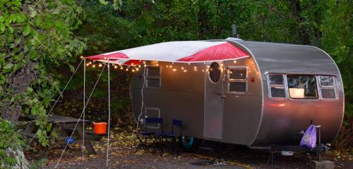 A vintage silver camper with a red and white awning, string lights, and two chairs outside in a wooded area.