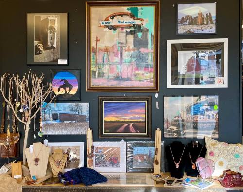 A display of artwork and handmade items on a black wall, featuring paintings, photographs, and jewelry.