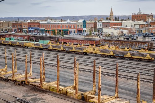 A view of a train yard with freight cars and a small town in the background, featuring buildings and a church steeple.