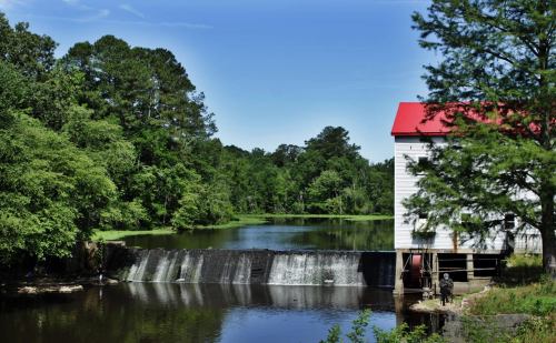 A serene scene of a white building with a red roof beside a waterfall, surrounded by lush green trees and a calm river.