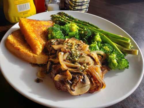 A plate featuring grilled steak topped with onions, served with asparagus, broccoli, and two slices of toasted bread.
