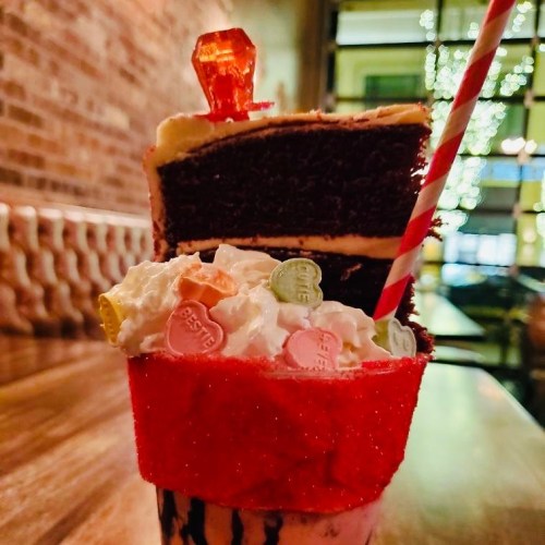 A tall dessert featuring a milkshake topped with whipped cream, candy, and a slice of chocolate cake with a red candy on top.