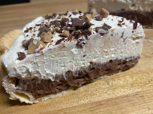 A slice of layered dessert with chocolate mousse, whipped cream, and chocolate shavings on top, resting on a wooden surface.