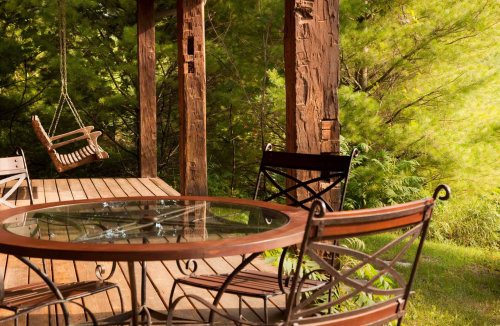 A wooden porch with a glass table and chairs, featuring a swing in a lush green setting.