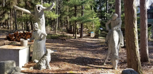 Two concrete sculptures in a wooded area: one of a man on a horse and another of a figure with outstretched arms, with a dog nearby.