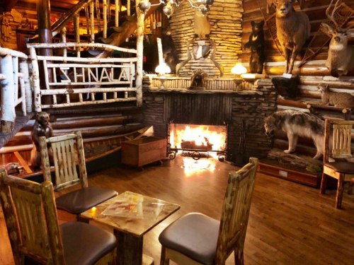 Cozy lodge interior with wooden furniture, a stone fireplace, and mounted animal trophies on the walls.