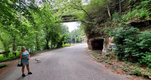 A woman stands by a tree-lined road with a small dog, near a stone tunnel and a bridge overhead.