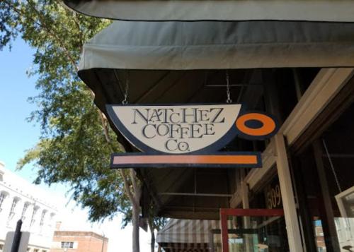 Sign for Natchez Coffee Co. hanging outside a café, with a blue and orange design against a sunny backdrop.