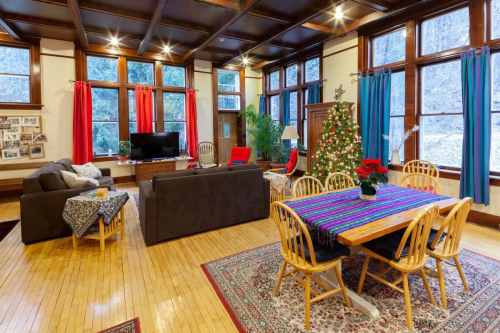 Cozy living room with a Christmas tree, colorful curtains, a dining table, and large windows letting in natural light.