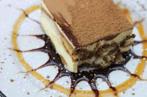 A slice of tiramisu on a plate, drizzled with chocolate sauce and sprinkled with cocoa powder and powdered sugar.