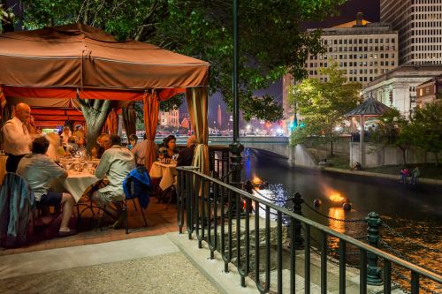 A riverside restaurant at night, with diners under tents and city lights reflecting on the water.