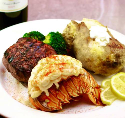 A plate featuring grilled steak, lobster tail, baked potato with sour cream, and steamed broccoli, garnished with lemon slices.
