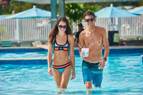 A man and woman walk through a pool, wearing swimwear and sunglasses, enjoying drinks on a sunny day.