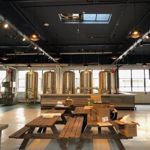 Interior of a brewery featuring wooden tables, brewing equipment, and large windows with natural light.