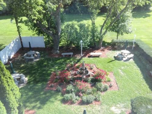 Aerial view of a landscaped garden with a fountain, flower beds, benches, and green grass surrounded by trees.
