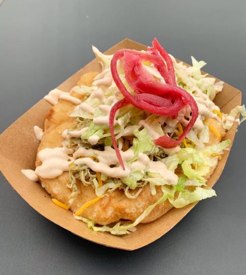 A loaded taco topped with shredded lettuce, cheese, pickled onions, and drizzled with sauce, served in a paper tray.