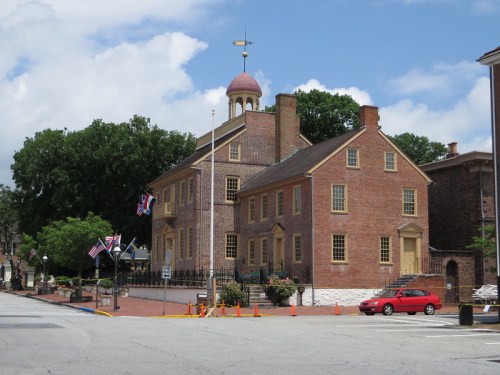 Historic brick building with a dome, surrounded by trees and flags, located on a sunny street.