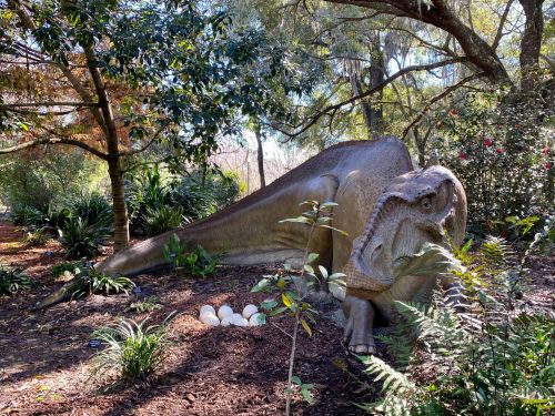 A lifelike dinosaur sculpture resting among greenery, with dinosaur eggs nearby in a natural setting.
