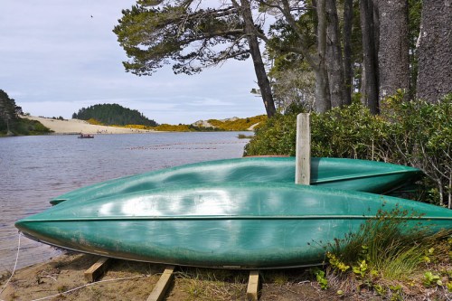 Two green kayaks are stacked on a sandy shore beside a calm lake, surrounded by trees and distant hills.
