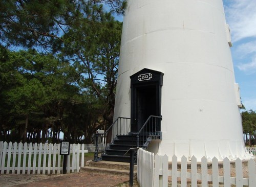 A white lighthouse with a black door and steps, surrounded by trees and a white picket fence.