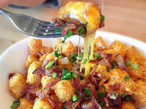 A fork lifting cheesy tater tots topped with bacon and green onions from a bowl.