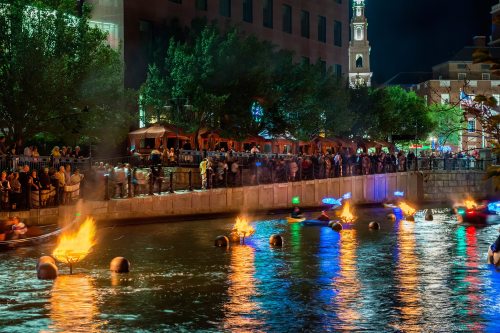 A vibrant night scene of a river with colorful lights, people gathered along the banks, and reflections in the water.