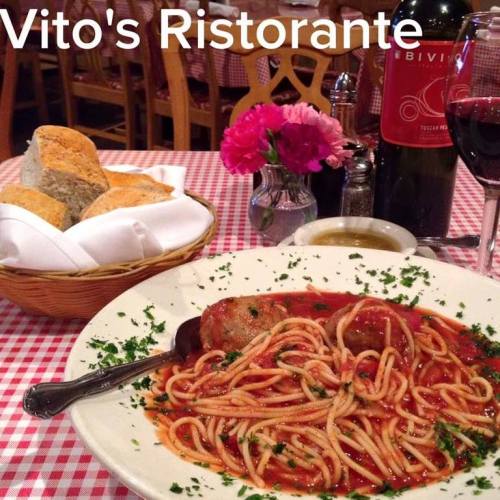 A plate of spaghetti with meatballs, garnished with parsley, alongside bread, wine, and a flower vase at Vito's Ristorante.