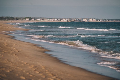 A serene beach scene with gentle waves lapping at the shore and distant houses lining the coastline.