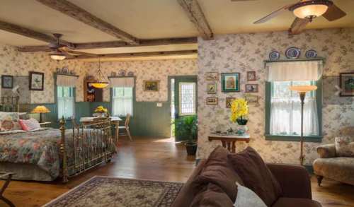 Cozy bedroom with floral decor, wooden beams, and a sitting area, featuring a bed, table, and plants.