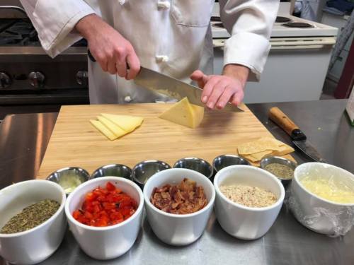 A chef slices cheese on a wooden cutting board, surrounded by bowls of diced tomatoes, bacon, herbs, and breadcrumbs.