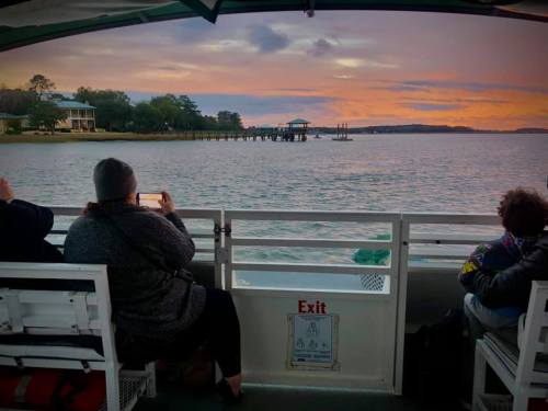 A sunset view from a boat, with passengers sitting and taking photos of the water and pier in the distance.