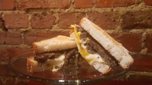 A close-up of a sandwich with melted cheese, beef patty, and egg, served on a glass plate against a brick wall.