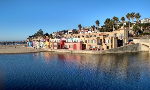 Colorful beach houses line the shore, reflecting in calm water under a clear blue sky. Palm trees dot the landscape.
