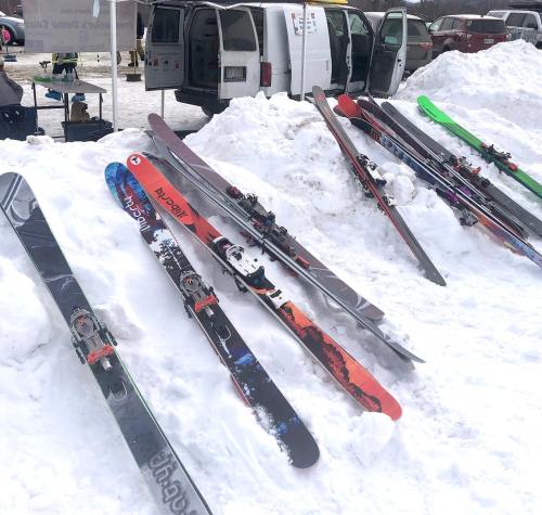 A row of colorful skis resting on a snowy surface, with a van and equipment in the background.