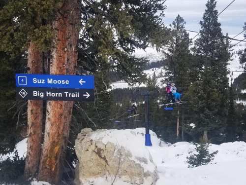 Skiers on a chairlift with signs for "Suz Moose" and "Big Horn Trail" amidst snowy trees and mountains.